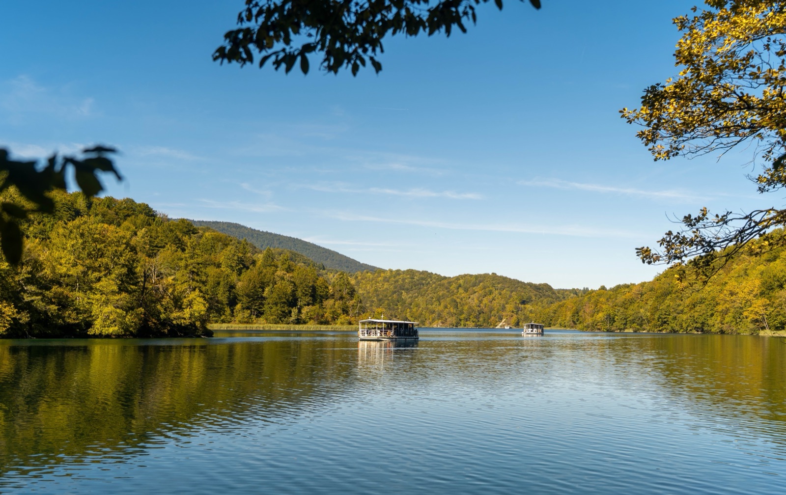 Two electric boats on a calm lake between wooded hills under a clear blue sky