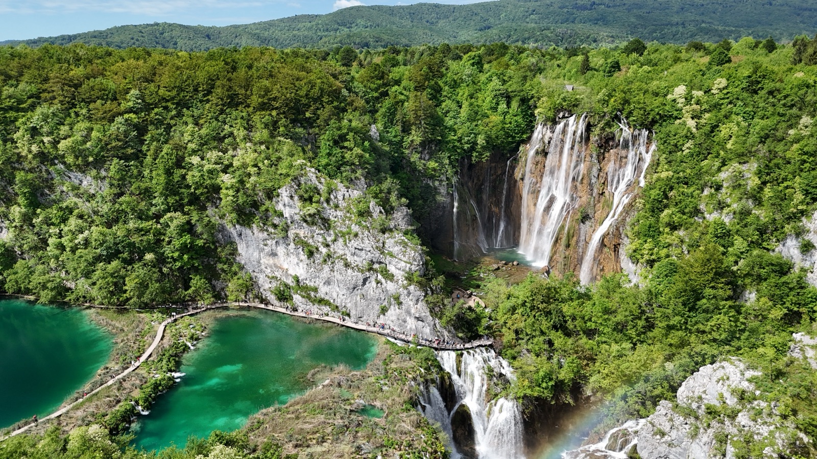 Aerial view of tall waterfalls dropping from a wooded cliff above turquoise lakes, with walkers on a wooden boardwalk